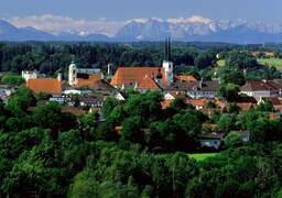 Altötting nahe dem 3-Sterne-S-Hotel und Landgasthof Happinger Hof beim Chiemsee Stadt Altötting mit Kirche nd mehreren Häusern, umgeben von Wäldern, dahinter Gebirge,